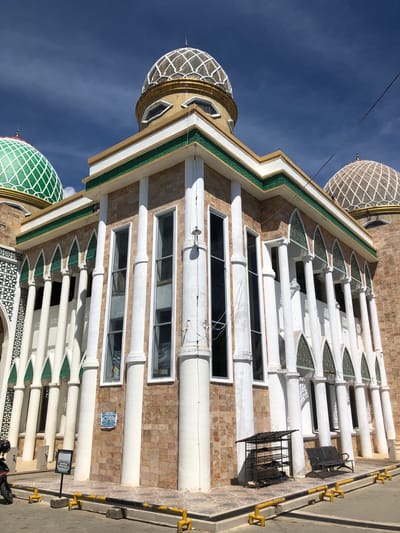The image depicts a mosque in Gampongjawa, Indonesia. The structure features a beige brick facade, rows of rectangular windows, and decorative white columns with pointed, triangular green accents. Two domes are visible: one with a green lattice and another with a white and gold lattice. The sky is blue with some clouds. A yellow and black bollard runs along the right edge of the image. A black metal rack and a sign are also visible near the base of the building.