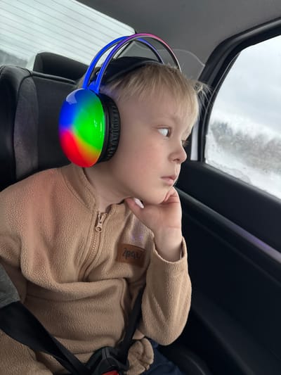 A young, fair-skinned child with blond hair sits in a car seat. The child is wearing a beige fleece jacket with a zipper and a logo patch on the upper left side. The child's left hand is raised to their chin, and they are gazing upwards with a thoughtful expression. The child is wearing headphones that feature rainbow-colored lights on the ear cups and blue lights on the headband. The car seat is secured with a black seatbelt. Through the car window, a snowy or icy landscape is visible.

There is no evidence to support the claim that this image was taken in the city of Samolet, Russia.