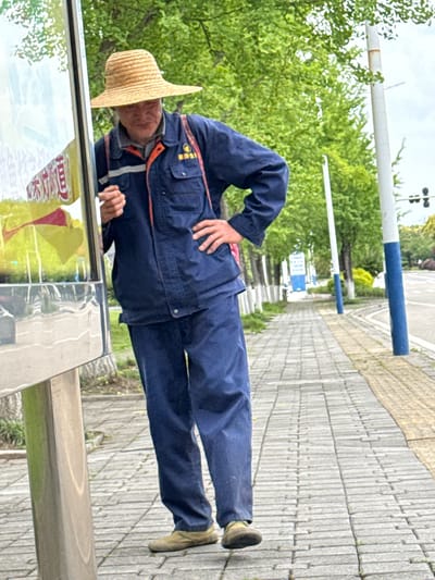 A man in blue work clothes and a straw hat leans over a bus stop advertisement in Unknown, China. He has his right hand on his hip and appears to be examining something in his left hand. The bus stop is located on a paved sidewalk lined with trees and blue and white posts.