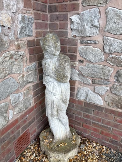 A pale stone statue of a reclining figure, likely an angel or similar allegorical figure, rests on a walkway constructed of red bricks alternating with sections of irregularly shaped grey stones. A stone plinth supporting the figure's lower body is surrounded by small pebbles and debris. The location appears to be outdoors, possibly in a garden or courtyard setting within the city of Shrewsbury, United Kingdom. A small drainage grate is visible near the base of the plinth.