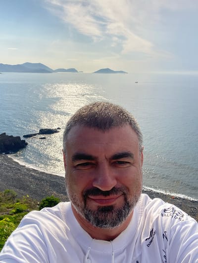 A man with a beard and short graying hair takes a selfie with a scenic coastal view in the background. He is wearing a white t-shirt with a Balenciaga logo visible on the shoulder and sleeve. The background features the ocean, a rocky coastline with dark pebbles, and distant islands under a partly cloudy sky. The sun is reflecting brightly on the water, creating a shimmering effect. Green vegetation can be seen on the left side of the image. The location is identified as the Nam Natural Heritage Area Trang, Vietnam.
