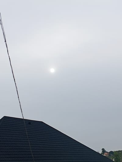 This is a full-frame, eye-level photograph taken outdoors in Ashata, Nigeria. The image features a dark, textured roof in the foreground, angled from the bottom left corner upwards towards the right. The roof appears to be made of asphalt shingles. A single, thin black wire runs diagonally from the top left edge of the frame down towards the roof. In the upper portion of the image, a bright, diffused sun is visible through a hazy, overcast sky. The sky occupies the majority of the frame, creating a muted and light-filled atmosphere. In the far background, on the right side of the frame, a few indistinct residential structures and some green foliage can be seen. There are no people or animals present in the scene. The overall impression is one of a cloudy day with the sun partially obscured.