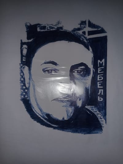 The image is a blue ink drawing on a textured white surface, possibly paper or a canvas. The drawing depicts a man's face, rendered in a sketch-like style with visible lines and shading. The man is shown from the shoulders up, with his head turned slightly to the right. His eyes are open, and he has a neutral expression. There are some abstract shapes or scribbles above and to the left of his head, as well as a vertical block of text to the right of his face. The text appears to be in Cyrillic script and reads "МЕБЕЛЬ," which translates to "FURNITURE" in English. The drawing is slightly abstract, with some areas of the face less defined than others. The lighting in the image suggests it was taken indoors, with a bright light source reflecting off the surface. There are no discernible elements that would place this drawing in a specific city, such as Gulbag, Uzbekistan. The image focuses solely on the artwork itself.