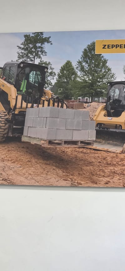 A close-up, slightly angled shot shows a construction scene with two yellow Caterpillar skid-steer loaders and a stack of concrete blocks. The loader in the foreground is to the left, with its forks holding a pallet of concrete blocks, and the loader in the background is to the right, with its front end slightly obscured by the blocks. The ground is covered in dirt and sand, and there are trees and buildings in the background. A yellow sign with the word "ZEPPEL" is visible in the upper right corner. The image does not provide any information about the city of Almalyk, Uzbekistan.