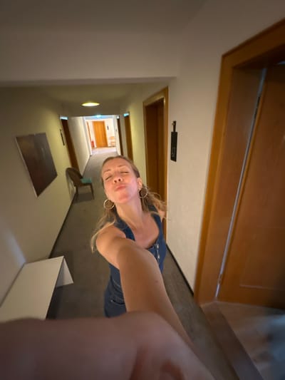 A woman with blonde hair is taking a selfie in a hotel hallway. She is wearing a blue denim dress and large hoop earrings. She is puckering her lips as she takes the photo, and her arm is extended towards the camera. The hallway is lined with doors and a carpeted floor. A framed picture hangs on the wall to the left, and a chair is visible further down the hall. The scene appears to be a hotel in St. Märgen, Germany, based on the provided information.