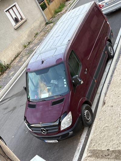 An overhead, slightly angled, medium shot captures a parked, dark red Mercedes-Benz Sprinter van on a paved street in Anton, Bulgaria. The van is positioned parallel to a raised concrete curb with double white painted lines indicating a parking area. To the left of the van, a building with a light-colored, textured facade and a window with white curtains is visible. A small amount of overgrown vegetation lines the base of the building and extends towards the street. The van's windshield reflects a pale light, and a person's blonde hair is visible through the glass. The license plate of the van reads "EH1116MA". Behind the Sprinter, a silver car is partially visible, with a license plate that appears to read "CB 0380 MX". The overall environment suggests a residential or commercial street setting.