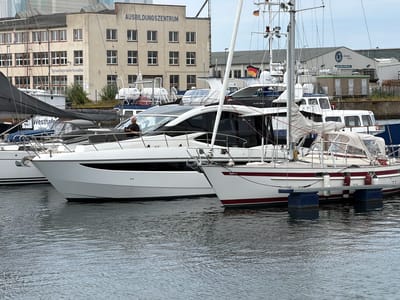 This is a medium shot of several yachts docked in the harbor of Wismar, Germany, on a cloudy day. In the background, a light-colored building with "AUSBILDUNGSZENTRUM" written across its facade stands out. To the left of the building, a smaller sign reads "Schweißtechnische Kursstätte". Several boats are visible in the foreground. The largest is a white yacht with a black stripe and a dark, tinted canopy. A man is sitting on the deck of this yacht, looking at his phone. To the right of this yacht is another white yacht with red stripes. Its sail is covered with a light-colored tarp, and it is tethered to a blue floating dock. In the background, other boats and industrial buildings are partially visible. The water is choppy with ripples reflecting the overcast sky.