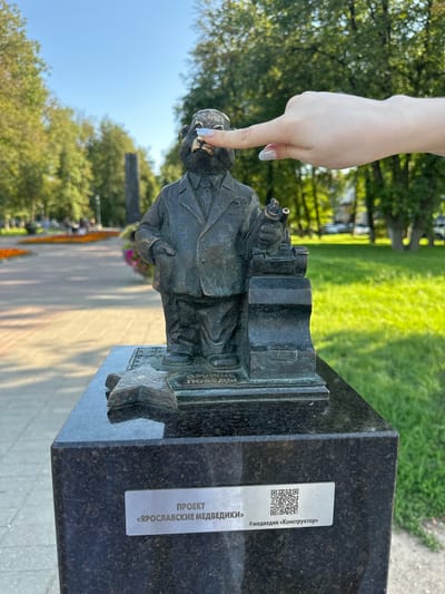 A bronze statue of a bear wearing a suit and tie stands on a black granite pedestal in a park. The bear is positioned next to a large mechanical contraption, possibly a printing press or a sewing machine, and is holding a spool of thread. A woman's hand is reaching out and gently touching the bear's nose with her index finger. The statue is located in Yazvitsevo, Russia, as indicated by the plaque on the pedestal. The background shows a sunny park with green grass, trees, and flower beds. The plaque on the pedestal reads "ПРОЕКТ «ЯРОСЛАВСКИЕ МЕДВЕДИКИ»" and "#медведик «Конструктор»".