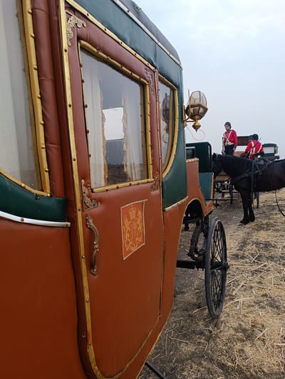 The image depicts an outdoor scene in Kottalapalli, India, featuring an ornate horse-drawn carriage in the foreground. The carriage is predominantly brown-orange with dark green upper sections and extensive gold trim. It has a large window with white curtains visible inside, an ornate gold door handle, and a gold-colored lantern mounted on its side. A rectangular crest, identified as the Royal Coat of Arms of the United Kingdom, is affixed to the carriage body. A black spoked wheel is partially visible at the bottom.
In the midground, two individuals in historical military-style uniforms are present. They wear red tunics with white cross belts and dark headwear. One individual stands upright, holding a long firearm vertically. The second individual is partially obscured, seated or stooping. A dark-colored horse, harnessed, stands between the primary carriage and a simpler, dark-colored cart in the background. The ground is covered with dry, light-colored straw or stubble over dark soil. The sky above is overcast and pale gray. A third person, partially visible in white clothing, is further back on the right.