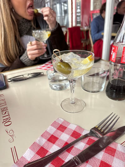 The image captures a dining scene, likely at a bistro in Bayonne, France, as suggested by the visible "AU P'TI BISTRO" inscription on the table. A person is holding a cocktail glass with ice and a lemon slice, skewering an olive with a cocktail pick, and positioning it towards their mouth. There is a Coca-Cola bottle and empty glass next to the cocktail glass. A fork and knife rest on a red and white checkered napkin. Another person is partially visible in the background. A pair of glasses and a cell phone are also visible on the table.
