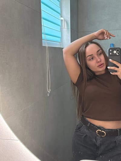 A young woman with long, brown hair and light skin is taking a selfie in a bathroom. She is wearing a brown, cropped t-shirt and dark, high-waisted jeans with a brown belt. Her left arm is raised behind her head, and she is looking at the camera with a neutral expression. In the background, a blue-striped window blind is visible, and the walls are covered in large, grey tiles. The scene is lit by a bright light source, likely from above. There is no information in the image to suggest the location is Kamyennoye, Russia.