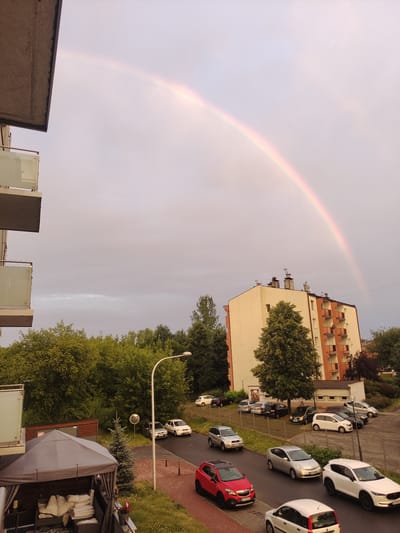 An aerial shot captures a cityscape with a prominent rainbow arching across a pale, cloudy sky. In the foreground, a portion of a modern apartment building's balcony railing and concrete overhang are visible on the left. Below, a gazebo with white cushions and furniture is set up on a green lawn. Beyond the gazebo, a street with several parked cars and a few moving vehicles is depicted. A taller, light-colored apartment building with orange accents stands to the right of the frame, framed by lush green trees. The scene is set in the city of Częstochowa, Poland.