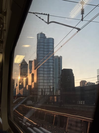 The image displays an exterior urban landscape viewed through the window of a train. The perspective is from inside the train, with the window frame, a portion of the interior wall, and ceiling visible on the left and top edges. The location is Philadelphia, United States of America, specifically looking east towards Center City from an elevated position over train tracks, likely near 30th Street Station.

The scene is illuminated by the low angle of the sun, indicating late afternoon or early evening. Golden sunlight reflects prominently off the glass facades of several buildings and the metal rails of the tracks. The sky is a light blue-gray.

In the midground, an extensive network of multiple parallel train tracks is visible, featuring ballast, wooden ties, and utility pipes running alongside. Overhead, a catenary system of electrical wires and support structures, including an insulator, partially obscures the sky.

The background is dominated by a dense concentration of high-rise buildings. The most prominent structure is 1818 Market Street, a tall skyscraper with a distinctive wavy, blue-tinted glass facade, centrally located. To its right stands 1919 Market Street, a residential tower with a lighter blue glass exterior. To the left of 1818 Market Street, the FMC Tower at Cira Centre South is visible, heavily reflecting the golden sunlight. Other commercial and residential buildings, varying in height and design, form the remainder of the skyline, with several exhibiting strong solar reflections.

Reflections of interior train lights, appearing as rectangular patterns, are superimposed on the window glass, along with faint reflections of other internal train elements. No individuals or moving vehicles are discernibly present in the external environment.