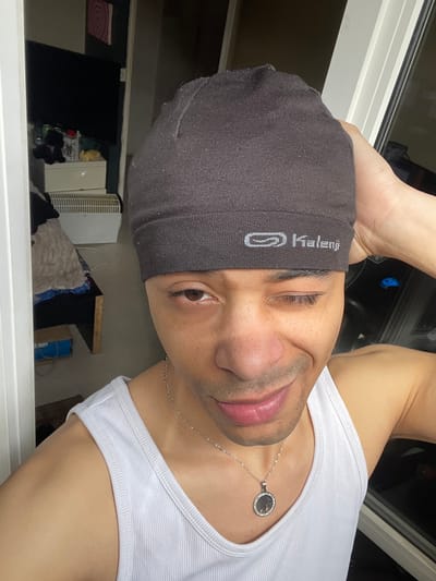 The image depicts a close-up, chest-up selfie of a male individual, located in Plaisir, France. The subject, who has a medium-dark skin tone, is wearing a dark gray ribbed beanie with the "Kalennji" logo visible on the front and a white ribbed tank top. A silver chain necklace with a circular pendant featuring a "tree of life" design, encircled by small clear stones, hangs around his neck. His right eye (viewer's left) is open and directed at the camera, while his left eye (viewer's right) is closed in a wink. He exhibits light facial hair and a slight smile. His right hand (viewer's left) is raised to the back of his head, with fingers visible gripping the beanie.

The background on the left, partially framed by a white door jamb, reveals a dimly lit interior space. A dark flat-screen television is mounted on a wall or positioned on a white two-drawer storage unit. On top of this unit, a black plush toy, a lighter-colored ceramic mug, and a small yellow plush object are present. Below the unit, a white horizontal radiator or heater is visible. A dark, possibly wooden, surface extends from the left, with a blue box resting on the floor beneath it. A pink/red circular abstract artwork is affixed to the wall above the television. The background on the right shows a window with white frames, reflecting a dark exterior. A faint blue reflection is discernible in the window glass behind the subject's right shoulder.