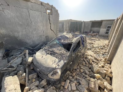 The image depicts a scene of extensive structural damage and debris in Spin Boldak, Afghanistan. A heavily damaged, dark-colored compact passenger vehicle, possibly a hatchback, is centrally positioned, partially buried under a large accumulation of rubble consisting primarily of broken bricks and concrete fragments. The vehicle's roof is crushed inward and filled with debris, its windshield is absent, and the front passenger side headlight is broken. A piece of blue fabric is draped over the driver's side roof rail.
To the left, a large, severely cracked concrete or mud-brick wall stands, featuring a prominent vertical fissure extending upwards from the ground to a small, high rectangular opening. The ground immediately surrounding the vehicle is entirely covered with construction debris. In the background, several additional structures are visible, appearing as concrete or mud-brick walls and buildings. A green metal gate is discernible in the far background. At least three motorcycles are parked on the dusty, uneven ground behind the damaged vehicle. No individuals are present in the scene. The environment is under clear, direct sunlight, indicating daytime conditions.