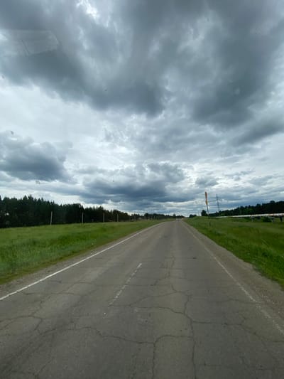 The image shows a two-lane asphalt road with a white line marking the edge. The road is cracked and appears to be in disrepair in some sections. On both sides of the road, there is a green grassy verge. To the left of the road, there is a dense line of dark green trees. To the right, the grassy verge continues, and further in the distance, there are some white pipes supported by structures, possibly an industrial pipeline. The sky is overcast with heavy, dark gray clouds, suggesting an impending storm or inclement weather. In the far distance, there are some indistinct shapes of buildings and a transmission tower. The scene appears to be a rural or semi-rural highway location.