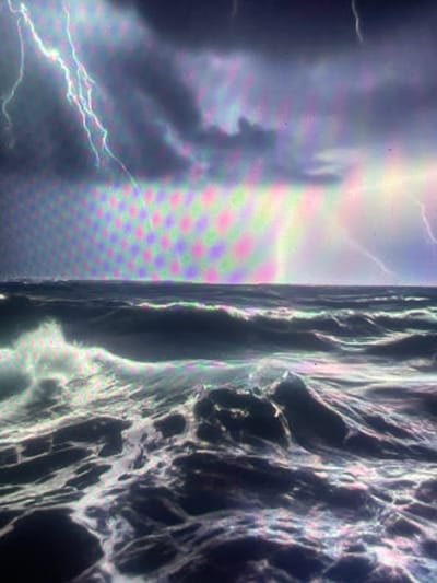 This image is a photograph of a digital screen displaying a stormy open ocean scene. The sky is dominated by dark, heavy clouds, with multiple bolts of lightning striking across the upper left quadrant and a luminous glow on the right horizon. The sea below is rough, characterized by large, dark waves and white crests of foam, particularly prominent in the foreground. A distinct, repeating grid pattern of multi-colored subpixels (magenta, green, blue, and cyan) is visible across the entire image, a characteristic artifact of photographing a digital display. The depicted environment is an open ocean under severe weather conditions, devoid of any human presence, specific man-made objects, or interactions. In the context of Zelenyi Hai, Ukraine, this image is an observation of a digital screen presenting this severe weather landscape.