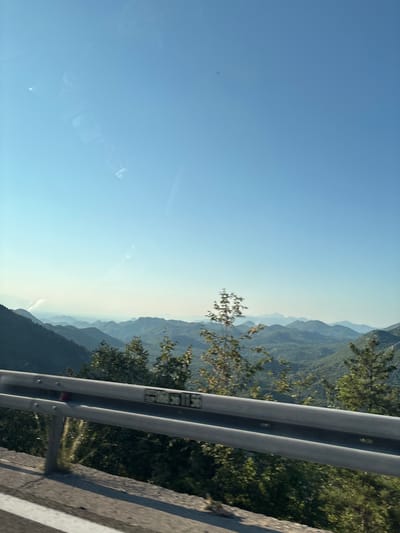 The image shows a mountainous landscape seen from a road with a guardrail in the foreground. The sky is clear and blue. Layers of mountains recede into the distance, with the closest mountains covered in lush green vegetation and the farther mountains appearing as hazy blue silhouettes. The guardrail is metallic and shows some signs of wear. A portion of the road with a white line is visible on the lower left. The environment suggests a scenic route in a mountainous region. This view is characteristic of the area around Dobrsko selo in Montenegro, known for its natural beauty and winding roads through the mountains.