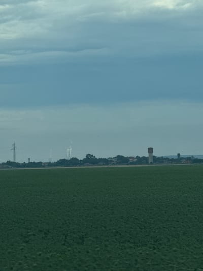 A wide shot of a rural landscape under a cloudy sky shows a vast, dark green field of crops in the foreground. In the mid-ground, a low-lying treeline separates the field from a village with red-roofed buildings. A tall, narrow water tower stands out in the village. To the left of the water tower, a tall electricity pylon is visible, and further left, a faint plume of smoke rises from a structure in the distance. The sky is a dull, overcast blue.