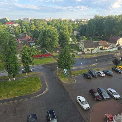 An aerial view captures a suburban street scene in Trykovka, Russia, under a partly cloudy sky. A wet asphalt road curves through the frame, bordered by patches of green grass and mature trees. Several houses with various roof designs, including red and grey shingles, are visible, interspersed with dense foliage and some taller apartment buildings in the distance.

A number of cars are parked or driving on the road and in parking areas. These include a white sedan on the left, a silver hatchback, two dark SUVs, a maroon wagon, a dark grey sedan, and a silver sedan parked in a row. Further down the road, a yellow car is visible. A few people are seen walking on a sidewalk next to a red fence. One of the houses is painted light green with a grey roof. A traffic sign with a yellow diamond shape is posted near an intersection. A green public information kiosk stands on a traffic island. The overall impression is a quiet residential area, likely after a rain shower.
