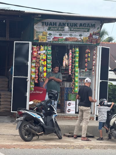 An exterior street-level, eye-level shot depicts a small, open-air convenience store, likely a "warung," in the city of Ajuen, Indonesia. A large, horizontal banner above the store entrance reads "TUAH ANEUK AGAM Jual B..." and "g Kelontong," indicating it sells various goods. The store itself is filled with shelves displaying numerous bagged snacks, beverages, and packaged food items.
In the foreground, a black and white scooter is parked. A man with dark hair and wearing a green t-shirt stands to the right of the scooter, facing the store and smiling. To the right of him, another man wearing a white baseball cap and a black t-shirt is facing away from the camera, seemingly interacting with the store's contents. A young boy in a striped shirt and shorts stands to the far right, holding onto the handlebars of a motorcycle. The ground surface is a mix of concrete and dirt. Stacked sacks of what appear to be cement or building materials are visible to the left of the store. The environment suggests a moderately busy, possibly humid, day with the store operating on a street.