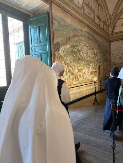 In the Palace of the Escorial, San Lorenzo De El Escorial, Spain, two people dressed as nuns stand in a room with a large mural on the wall. The first nun, in the foreground, is mostly obscured by a large white habit, with only a white wimple visible. The second nun, in the middle ground, wears a white wimple and a dark vest over a white shirt, with her back to the camera. A third person, a man in a black shirt and dark pants, stands further back in the room. The room has a wooden floor and a large mural depicting a historical scene covers the wall. A velvet rope barrier separates the visitors from the mural. A window with green shutters is visible on the left side of the image.