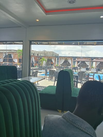 The image shows the interior of a restaurant located in Ujevwu, Nigeria (latitude: 5.471, longitude: 5.818). The restaurant is modern with white walls and a red accent strip on the ceiling. The large windows look out onto an outdoor seating area and a swimming pool. The restaurant is decorated with green velvet booths and a wooden table in the foreground. A person wearing a blue shirt is sitting in one of the booths, partially obscured by the plush back of the booth. The person is facing the window, looking out at the outdoor scene. The exterior view shows a variety of tables and chairs arranged around the pool, which is surrounded by palm trees and lush greenery. The overall scene appears to be upscale and modern, suggesting that the restaurant is a popular destination for dining and socializing.