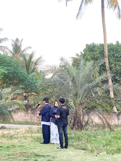 This media file shows a group of people posing outdoors, likely in a tropical or subtropical environment given the presence of palm trees and lush greenery. The scene appears to be a casual gathering.
The subjects include three individuals. Two are men dressed in black, one wearing a shirt with a prominent "M" logo on the back. The third person is a woman wearing a hijab and a dark blue outfit with white patterned pants. They are standing side-by-side on a grassy area, with palm trees and other foliage in the background. A brick wall is visible behind them.
The image is oriented vertically, suggesting it was taken with the camera held in portrait mode. The lighting suggests it is daytime, possibly late afternoon given the angle of light, though the sky is very bright. The weather appears to be fair.
The activity seems to be a photo opportunity, with the individuals looking towards the camera or slightly away from it. Their expressions are neutral, indicating a posed shot rather than a candid moment. The location context of Kembang Harum, Indonesia, aligns with the tropical vegetation depicted.
There is visible text on the back of one of the men's shirts: a stylized "M" with some additional lettering that is not clearly legible.