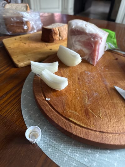 A medium shot captures a close-up of a rustic food preparation scene on a wooden table, likely in a kitchen or dining area in Krapivka, Russia. The primary focus is a well-worn round wooden cutting board, featuring several wedges of white onion and the tip of a knife blade. To the left of this board, a rectangular wooden board is partially visible, holding a block of what appears to be pork fat or lard (salo) with a pinkish layer of meat, still partially encased in clear plastic wrap. Further to the left, several slices of dark brown bread are visible, with some appearing to be in a clear plastic bag.
A light green placemat with a subtle white grid pattern is partially visible under the round cutting board and extending onto the wooden table, which displays a distinct wood grain. In the lower right corner of the frame, a small, white ridged plastic bottle cap lies on the placemat. The arrangement of ingredients suggests the preparation of a traditional snack or meal, possibly involving salo, onion, and bread, which are staples in Eastern European cuisine. The scene appears to be illuminated by ambient daytime light. There are no visible people or discernible text in the image.