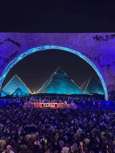 The image depicts the Great Pyramids of Giza in Egypt illuminated with blue light, seen behind a large semi-circular blue light display. A crowd of people is densely packed in front of the pyramids, facing them. A sign with the text "ZAMNA" in vertical letters is visible. The background is dark.