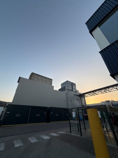 This is a wide-angle, low-angle, eye-level shot of an industrial building complex in Boxtel, Netherlands, under a clear, pale blue sky transitioning to a warm orange hue near the horizon, indicating sunrise or sunset. The structures are modern and industrial, featuring corrugated metal siding and large dark-colored doors with diamond-shaped patterns. One building has a distinctive sloped facade. Another section features a towering structure with multiple stacked units. A metallic walkway or bridge spans between buildings overhead. In the foreground, a paved area with crosswalk markings leads towards a covered waiting area with glass panels and a bright yellow protective barrier. The scene is devoid of significant human activity, with only faint reflections of people visible in the glass.