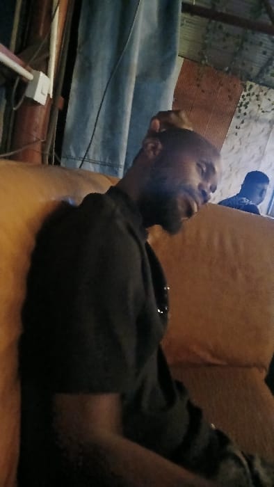 A man with a beard rests on an orange couch. He wears a black shirt. Another person wearing a blue shirt and a black hat is visible in the background. The surrounding area is cluttered with various objects, including what appears to be a blue tarp or sheet, some pipes or poles, and a white electrical outlet. The scene suggests an indoor environment, possibly a living room or recreational space.