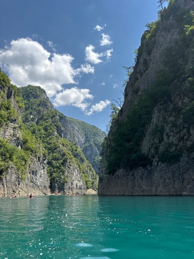 In the city of Bezuje, Montenegro, a serene turquoise lake stretches between towering, sheer cliffs draped with lush green vegetation. The water's surface is rippled, with small white bubbles dotting its expanse, hinting at a gentle current. In the distance, several small boats, occupied by individuals, navigate the tranquil waters. Above, a vibrant blue sky is punctuated by scattered white clouds, casting a dynamic play of light and shadow upon the majestic landscape.
