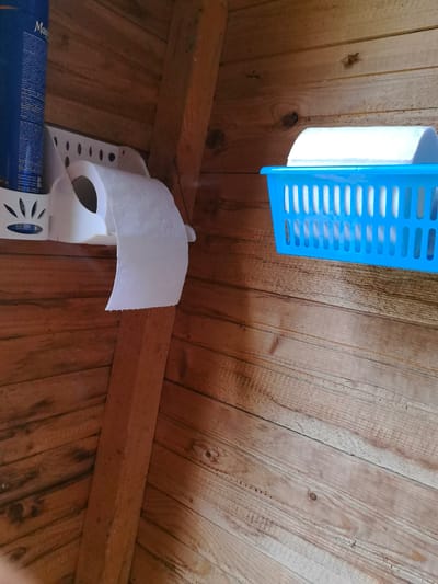 A toilet paper dispenser is mounted on a white bracket on a wooden wall. A roll of toilet paper is attached to the dispenser, with some of the paper unrolled. To the right, a blue plastic basket sits on the wall, holding more rolls of toilet paper. The walls and floor are made of rough, unfinished wood. The scene appears to be inside a rustic structure, possibly a shed or outhouse.