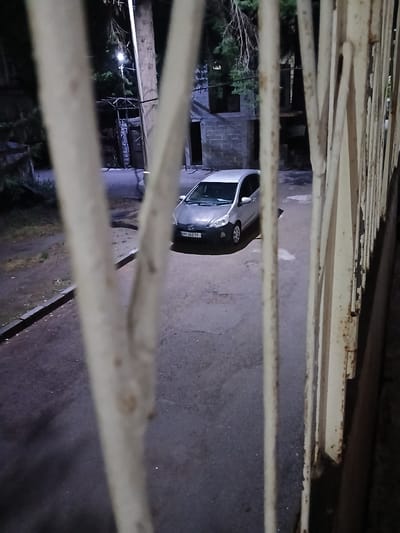 A silver hatchback car with license plate "MT 263 TY" is parked on a paved surface in front of a brick building. The car is viewed from behind a light-colored, rusty metal fence. Trees and branches are visible above the car and building. The scene appears to be an outdoor area, possibly a residential courtyard or parking lot, in the city of Chiatura, Georgia. The lighting suggests it is evening or night.