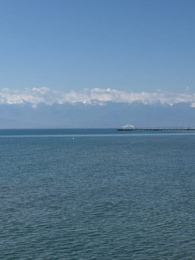 A wide shot captures a serene, vast body of blue water under a clear, pale blue sky. In the distance, a long, narrow pier extends from the horizon, featuring a white, domed structure at its end. Beyond the pier and water, a range of snow-capped mountains forms a majestic backdrop, their peaks softened by a thin layer of white clouds. The water's surface is gently rippled, and a few small, white objects, possibly buoys or birds, are scattered across it. The scene suggests a tranquil lakeside or coastal setting in Koshkol', Kyrgyzstan, likely during the daytime.
