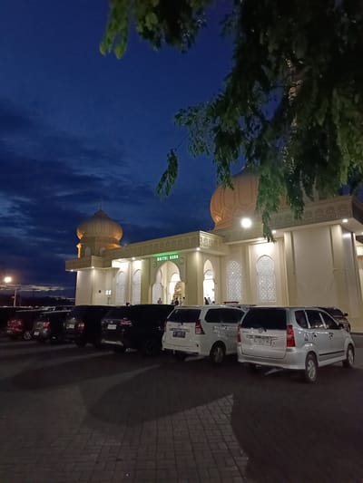 The image depicts a mosque, labeled "RAIYATUL MUDA," at dusk in Mee Tanjong Usi, Indonesia. The building features a prominent golden dome and intricate white latticework on its facade. A row of parked cars, including several white MPVs and dark SUVs, is visible in the foreground. The sky is a deep blue with scattered clouds, and the scene is illuminated by artificial lights. Some green foliage hangs into the frame from the top right. A few individuals can be seen near the entrance of the mosque.
