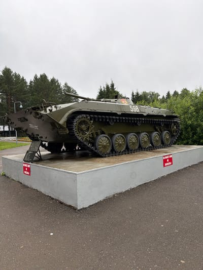 This media file shows a military tank on display in an outdoor setting. The tank is painted olive green and has the number "598" prominently displayed on its side. It is positioned on a raised concrete platform, which is part of a memorial or museum exhibit. Red signs with white lettering are attached to the platform, one reading "НА МАКЕТ БМП НЕ ЗАЛЕЗАТЬ!" which translates to "DO NOT CLIMB ON THE BMP MODEL!". The background features lush green trees and an overcast sky, suggesting it is daytime with potentially cloudy weather. The ground around the platform appears to be asphalt. The scene evokes a sense of historical commemoration or public display of military hardware. The presence of the signs indicates that the tank is an exhibit and visitors are expected not to interact with it physically by climbing on it. The tank itself appears to be well-maintained, possibly a static display for educational or remembrance purposes.