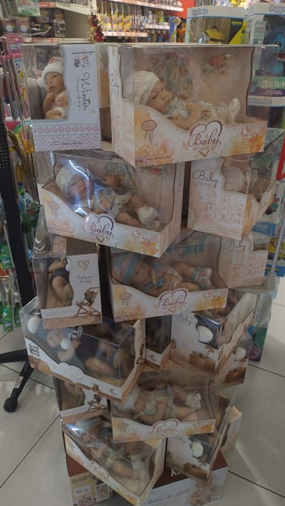 The image shows a display of boxed baby dolls in a retail environment, likely a toy store or department store. The dolls are packaged in clear plastic boxes with decorative cardboard inserts. Some of the boxes have text that reads "Baby" and "Grows up with you". The dolls appear to be various designs, some with hats and sleeping attire, and some with blankets. They are stacked in a somewhat disorganized manner on shelves. In the background, there are more shelves with various products, some of which are colorful toys. The flooring appears to be tiled. The lighting suggests an indoor setting. The presence of the dolls and packaging indicates a commercial space. The location provided is Pylaia Municipal Unit, Greece.
