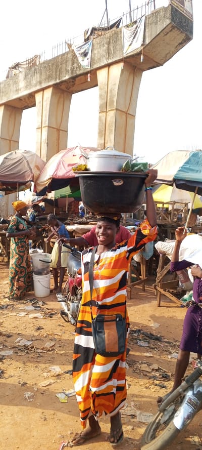 A smiling woman balances a large black tub filled with cooking pots and wrapped items on her head. She wears a striped orange, white, and black outfit with a small crossbody bag. The scene is a busy outdoor market under a concrete overpass. Numerous vendors with umbrellas and makeshift stalls fill the dirt ground scattered with litter. Other people are visible around her, including one wearing a yellow headscarf and another in a purple dress. A motorcycle is parked nearby.