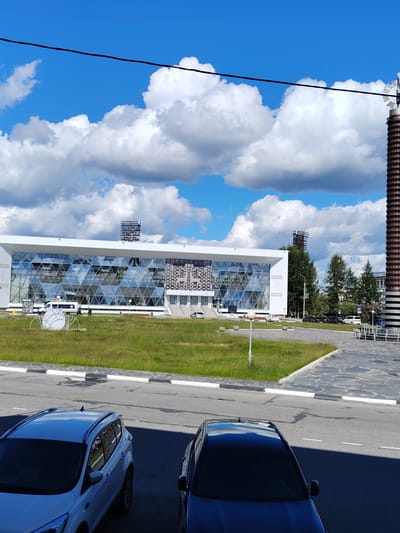 The image captures an outdoor scene in Olenegorsk, Russia, on a bright, cloudy day. In the foreground, two parked cars are visible: a white SUV on the left and a dark sedan on the right. Beyond the cars is a paved area with white lines, bordered by a grassy median. Further back, a modern building with a glass facade featuring a geometric pattern dominates the mid-ground. In front of the building, a white van and a few individuals are present, along with an abstract sculpture. The background includes trees, another building, and a tall, cylindrical structure. The sky is a vibrant blue, punctuated by large, fluffy white clouds.