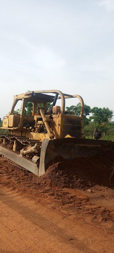A yellow, weathered D-8K bulldozer is the prominent subject in this outdoor scene, set in Otunade, Nigeria. The heavy machinery, a Caterpillar D8K model indicated by the "D-8K" text visible on its side, occupies the foreground and midground. It is situated on and surrounded by reddish-brown earth, with its large front blade lowered and pushing against a substantial pile of the same soil, suggesting recent land-clearing or earthmoving activity. The bulldozer shows clear signs of heavy use, with dirt, rust, and wear across its metallic frame, tracks, and exposed mechanical parts. An open cab, protected by a robust rollover structure, is visible, and a plastic jerry can along with other items are placed on a platform near the cab.

In the middle-right background, a man stands partially obscured by the bulldozer's blade and surrounding foliage. He wears a dark shirt and appears to be observing the scene. The background is characterized by dense green trees and vegetation, indicative of a lush, possibly rural, landscape. The sky above is bright and appears overcast or hazy, diffusing the light evenly across the scene. The overall impression is that of a construction or development site in a tropical environment, captured during the daytime.
