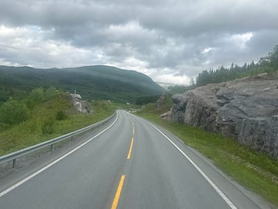 This media file captures a forward-facing view from a vehicle traveling on a paved, two-lane road in a mountainous, rural setting. The road itself is gray asphalt, marked with a broken yellow center line and a dashed white line on the right shoulder. A metal guardrail runs along the left side of the road.
The surrounding landscape is dominated by lush green vegetation, including trees and grass. To the left, the terrain slopes upwards, covered in dense foliage and some rocky outcrops. To the right, a steep, rocky cliff face rises alongside the road, composed of large, gray stones. In the distance, rolling hills and mountains are visible, cloaked in a layer of green forest. The sky is overcast with heavy, gray clouds, suggesting cool or impending rainy weather. The overall lighting is dim and diffused, indicating it is likely daytime, but not sunny.
There are no people visible in the media. A few vehicles can be seen in the distance on the road. A small white building is faintly visible nestled amongst the trees on the left side of the road in the mid-ground. A white and red marker post is visible on a rocky outcropping on the left.
The scene evokes a sense of travel through a remote, natural environment. The cloudy weather and imposing rock formations contribute to a slightly dramatic or moody atmosphere. The location context suggests this is Bjøllånes, Norway, consistent with the mountainous terrain and vegetation. There is no visible text in the media file.