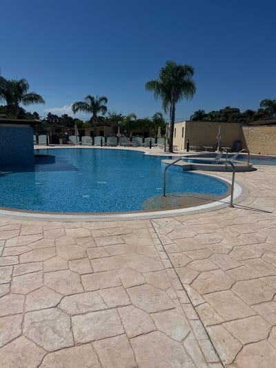 The image depicts an outdoor swimming pool area on a clear, sunny day. The pool is filled with bright blue water and surrounded by a light-colored, irregularly shaped paver patio. Palm trees are visible in the background, along with some buildings and lounge chairs. The overall scene suggests a resort or leisure facility. The specific location in Otranto, Italy, is not identifiable from the image alone, but the environment is consistent with a coastal town in southern Italy. There are no people visible in the image. The pool has a tiled section and a small jacuzzi area with water jets.