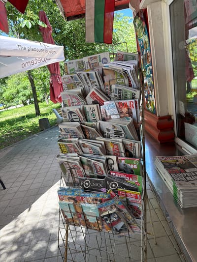 The image shows a newsstand in Chirpan, Bulgaria. The newsstand is made of a metal wire rack and contains a variety of Bulgarian newspapers and magazines. A Bulgarian flag is visible on the left side of the frame, adjacent to a white banner with text in Bulgarian. A sidewalk made of gray tiles is present to the right of the newsstand. The scene is outdoors on a sunny day, as indicated by the shadows and the bright green trees visible in the background. The newsstand is adjacent to a building with a window above and to the left.