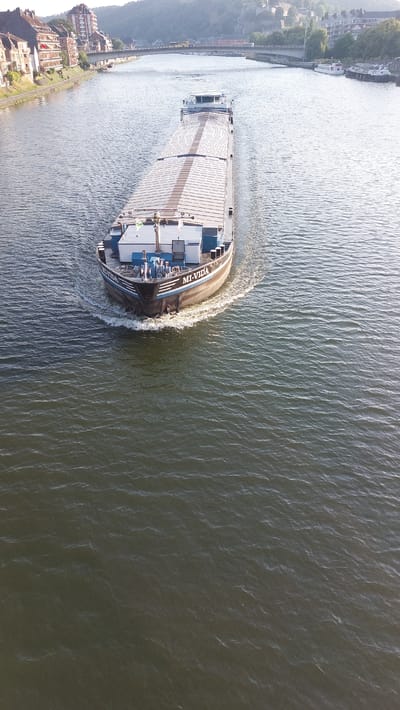 This is an aerial, high-angle drone shot of a barge navigating a river. The barge, with a flat, long deck covered by a tarp, is moving from the left to the right of the frame, creating a wake behind it. The barge appears to be a cargo vessel, likely transporting goods. On the stern of the barge, there is a small superstructure with windows and blue railings. Visible text on the side of the barge reads "MU-VIDA" and another line of text that is partially obscured.

The river occupies the majority of the frame, with its surface rippled by the movement of the barge and the breeze. The water is a murky green-brown color. To the left of the river, there is a shoreline with a towpath or road running along it. Beyond the path, there are buildings and a hillside densely covered with trees, suggesting an urban or semi-urban setting nestled within a natural landscape. The buildings on the left appear to be residential or commercial, with brick facades and multiple stories. The hillside is steep and verdant, with some structures visible even on its slopes.

The sunlight is bright, casting reflections on the water and illuminating the scene, indicating it is daytime. The sun appears to be low in the sky, possibly late afternoon or early morning, given the long shadows and the shimmering light on the water. The weather seems to be clear and fair.

The overall scene depicts a moment of transit for the barge, moving through a picturesque setting where developed land meets a natural river environment. The drone's perspective offers a unique, top-down view of the barge and its surroundings.