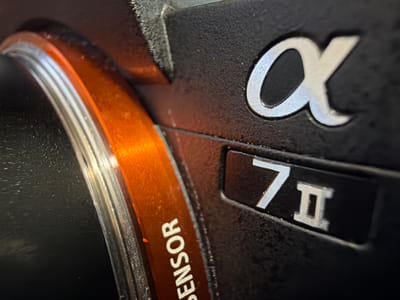The image is a close-up, macro photograph of the exterior of a black digital camera, likely a Sony Alpha series, given the prominent "α" logo. The camera body has a textured surface. To the left, there is an orange ring with the word "SENSOR" printed vertically in white. Further to the right, a black rectangular badge displays the text "7II" in white, indicating the camera model. The lighting suggests an indoor setting, with a warm orange light reflecting off the orange ring and a cooler, possibly bluish light reflecting off the silver lettering of the "α" and "7II" badges. The overall focus is on the branding and specific details of the camera's construction, with no visible environmental context or location information in the image itself. There are no people or actions depicted.