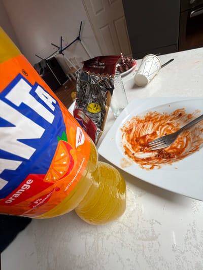 This image depicts an indoor scene in Wirges, Germany, likely a kitchen or dining area, viewed from a slightly elevated, oblique angle. The foreground is dominated by a large, clear plastic bottle of Fanta orange-flavored soda, partially shown and tilted, revealing its orange and blue label with "FANTA orange," "3% Orangensaft," and "mit Orangen-Geschmack" printed in German. The bottle contains a bright orange liquid.
On a light-colored, mottled countertop or table surface, several items are present. To the right of the Fanta bottle, a rectangular white plate holds residual red sauce, consistent with tomato or Bolognese sauce, along with a few small dark particulates. A silver-colored metal fork rests within the sauce on the plate. Behind the plate stands a clear, empty drinking glass. Further to the right and slightly behind, an open, metallic silver bag of sunflower seeds is visible, bearing a yellow circular sticker with "1€" (1 Euro). Next to the seed bag, a small white plate contains a partially consumed dessert item, appearing to be chocolate cake with red frosting or jam and a small pink decorative element. A white paper cup with a geometric pattern lies on its side near the cake, with a black spoon visible beside it.
In the blurred background, a dark piece of furniture or an appliance is visible to the left. A white interior door is partially discernable, and a blue and white clothes drying rack stands in the far background. The wall is light-colored. No individuals are present in the scene.
