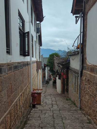 The image depicts a narrow, stone-paved alleyway in Gucheng District, China, flanked by traditional buildings. The buildings on either side feature white plastered walls, stone foundations, and dark tiled roofs with decorative eaves. The building on the left has several windows with dark wooden frames and one window is partially open. A white pipe runs vertically down the exterior of this building, attached to the stone wall. A red fire hydrant stands on the right side of the alleyway, next to a small wooden structure. The alleyway slopes downwards into the distance, where more buildings with red lanterns hanging from them are visible, along with some greenery. The sky is overcast with hints of mountains in the background.