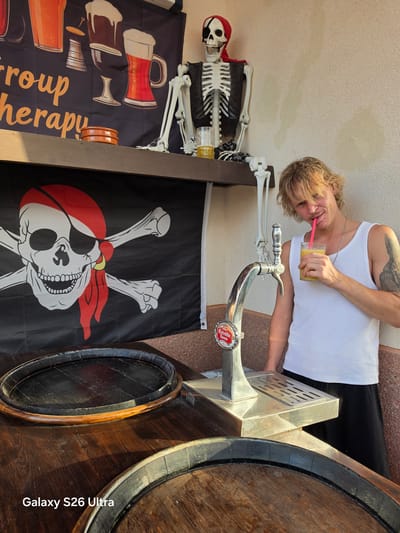 In the city of Llucmajor, Spain, a young man with blonde, wavy hair and a light mustache is leaning against a wall, drinking a yellow beverage through a pink straw from a clear glass. He is wearing a white tank top and black pants. To his left, a metal beer tap with a red and white logo is mounted on a wooden counter. Behind him, a skeleton prop wearing a red bandana and a black vest is seated, with a glass of amber liquid beside it. A black flag with a white skull and crossbones design is hanging behind the beer tap and skeleton. In the foreground, two wooden barrel rims are visible on a wooden surface. A banner with illustrations of various beers and the text "roup therapy" is displayed above the pirate flag.
